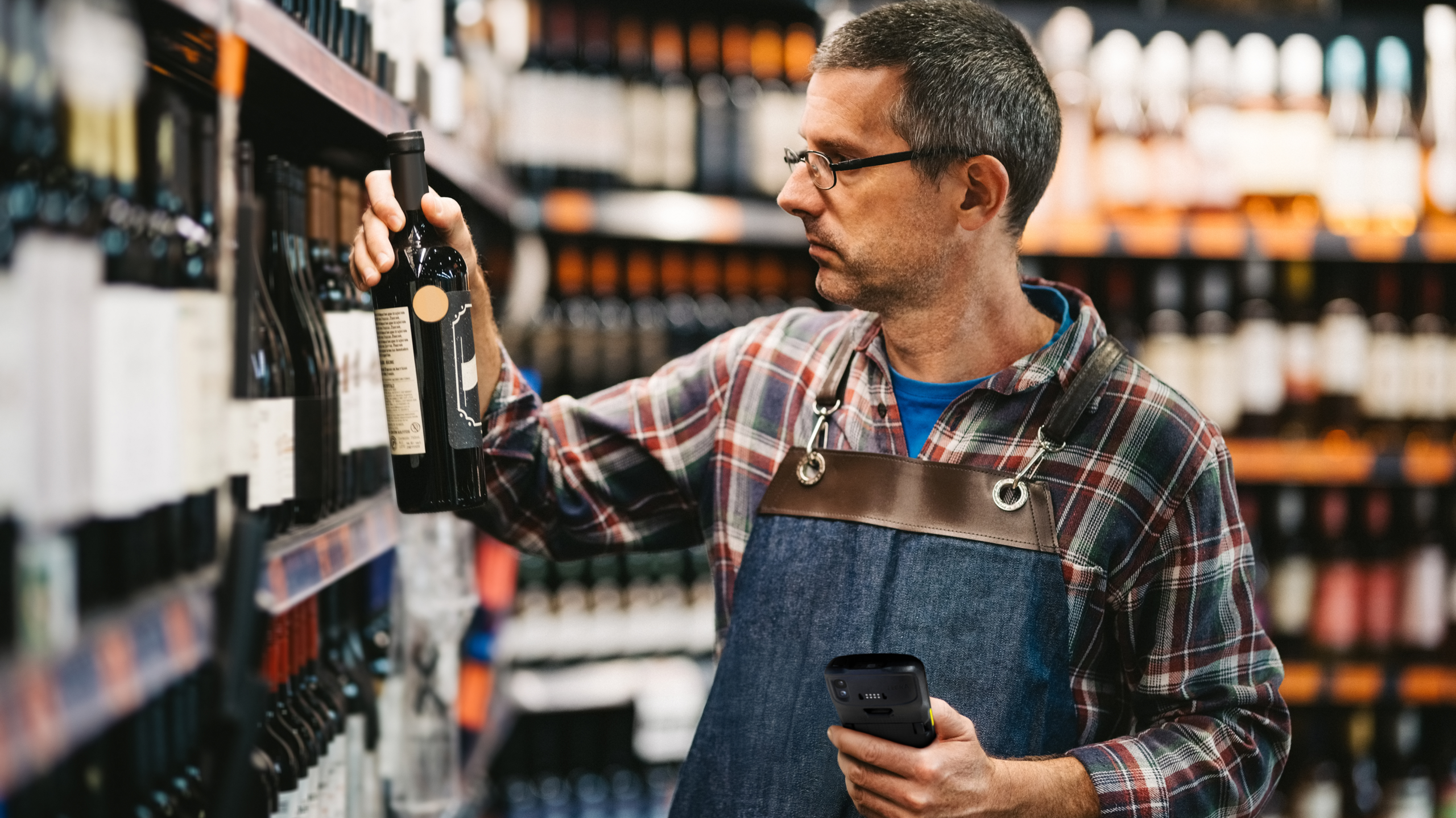 Retail employee scans a wine bottle in store aisle, checking product details while organizing inventory